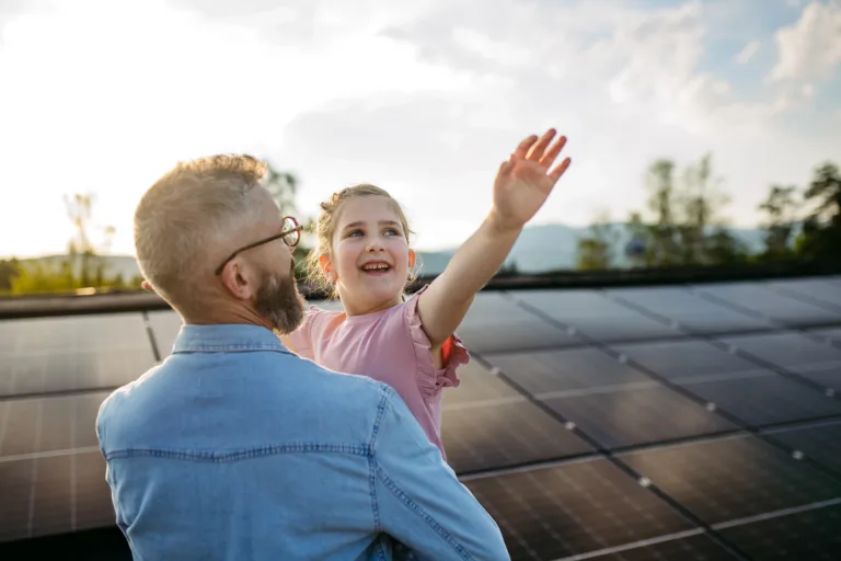 Adult holding a smiling child in front of rooftop solar panels.